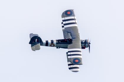 The Fairey Swordfish biplane in Royal Navy colours with black and white D-Day invasion stripes, banking against a pale sky with two crew visible in the open cockpit