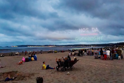 Drones spelling Nos Da above Goodnight over Swansea Bay, with crowds watching from the beach at dusk