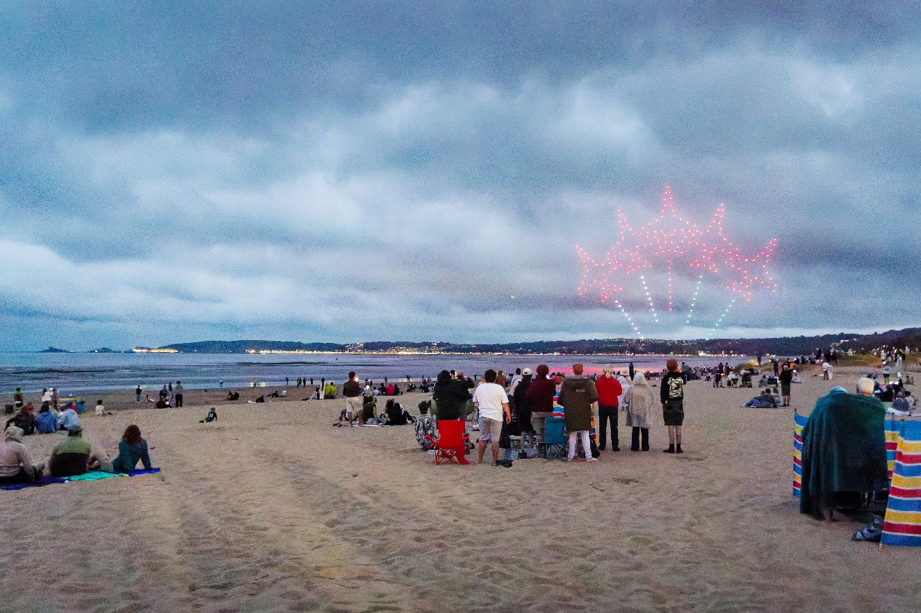 A drone formation of red aircraft lit above Swansea Bay at dusk, with crowds watching from the beach