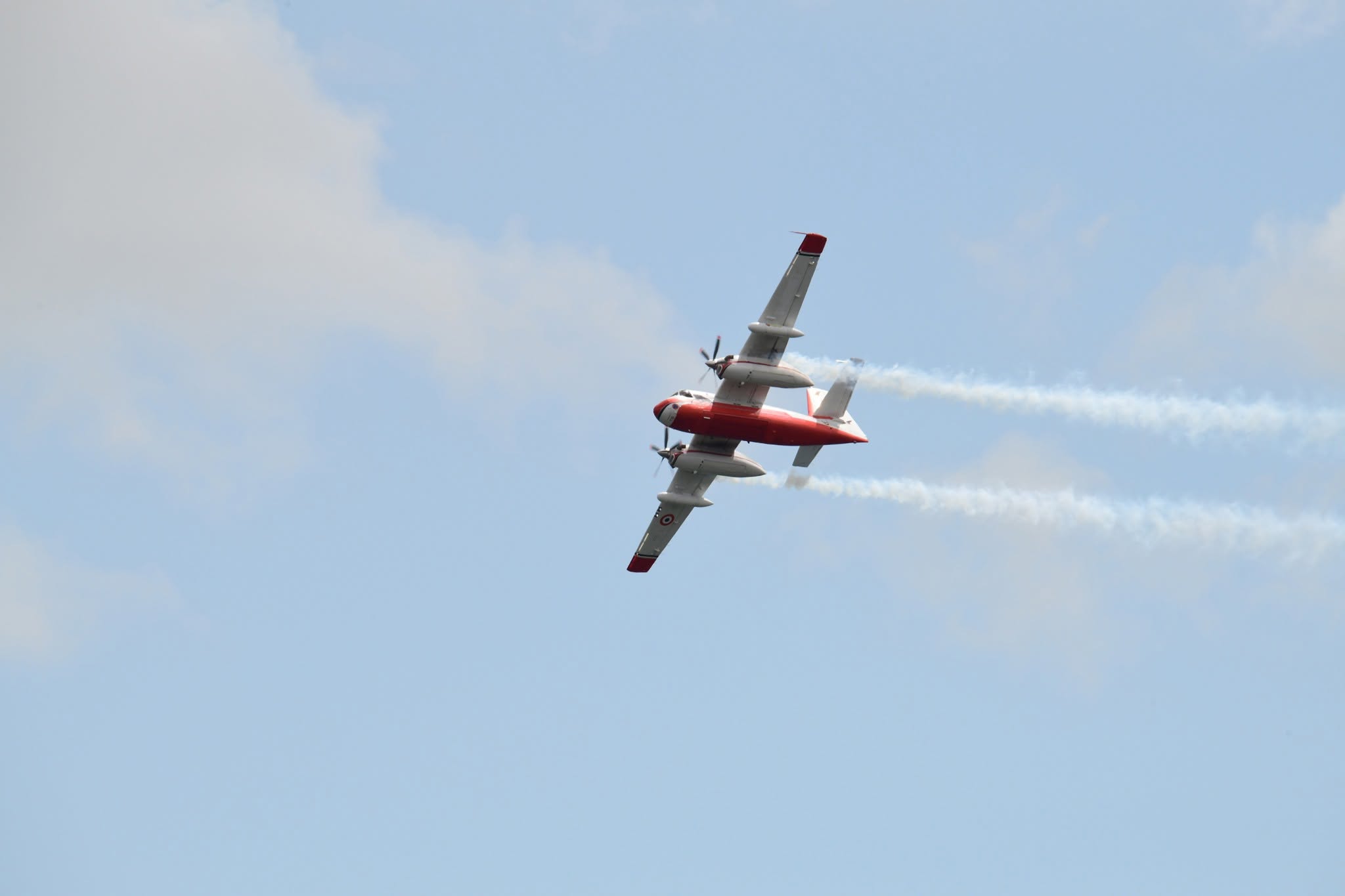 A red and white Firecat water bomber in French markings flying level with two white smoke trails