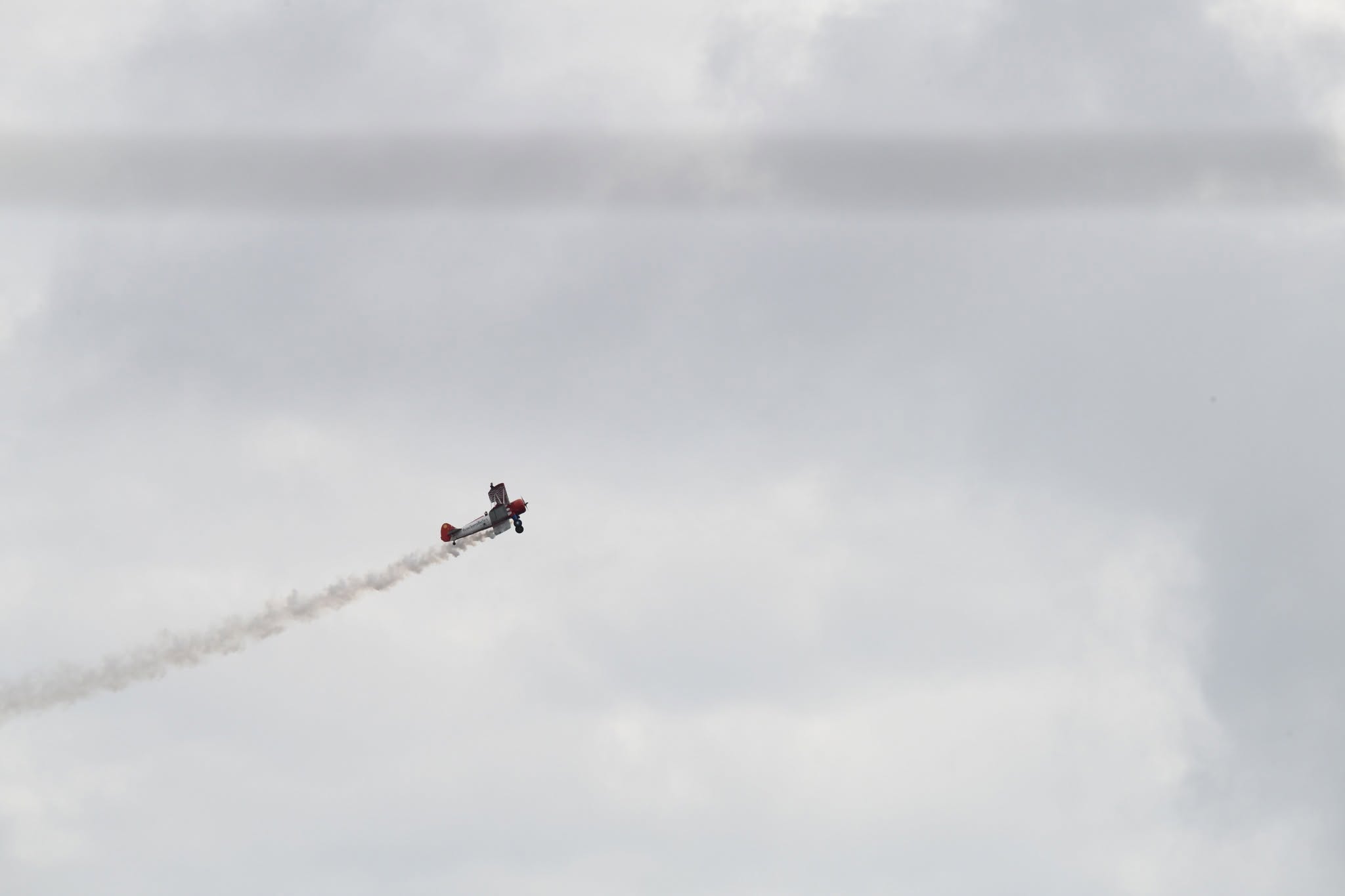 A Stearman biplane climbing away with a long white smoke trail behind it