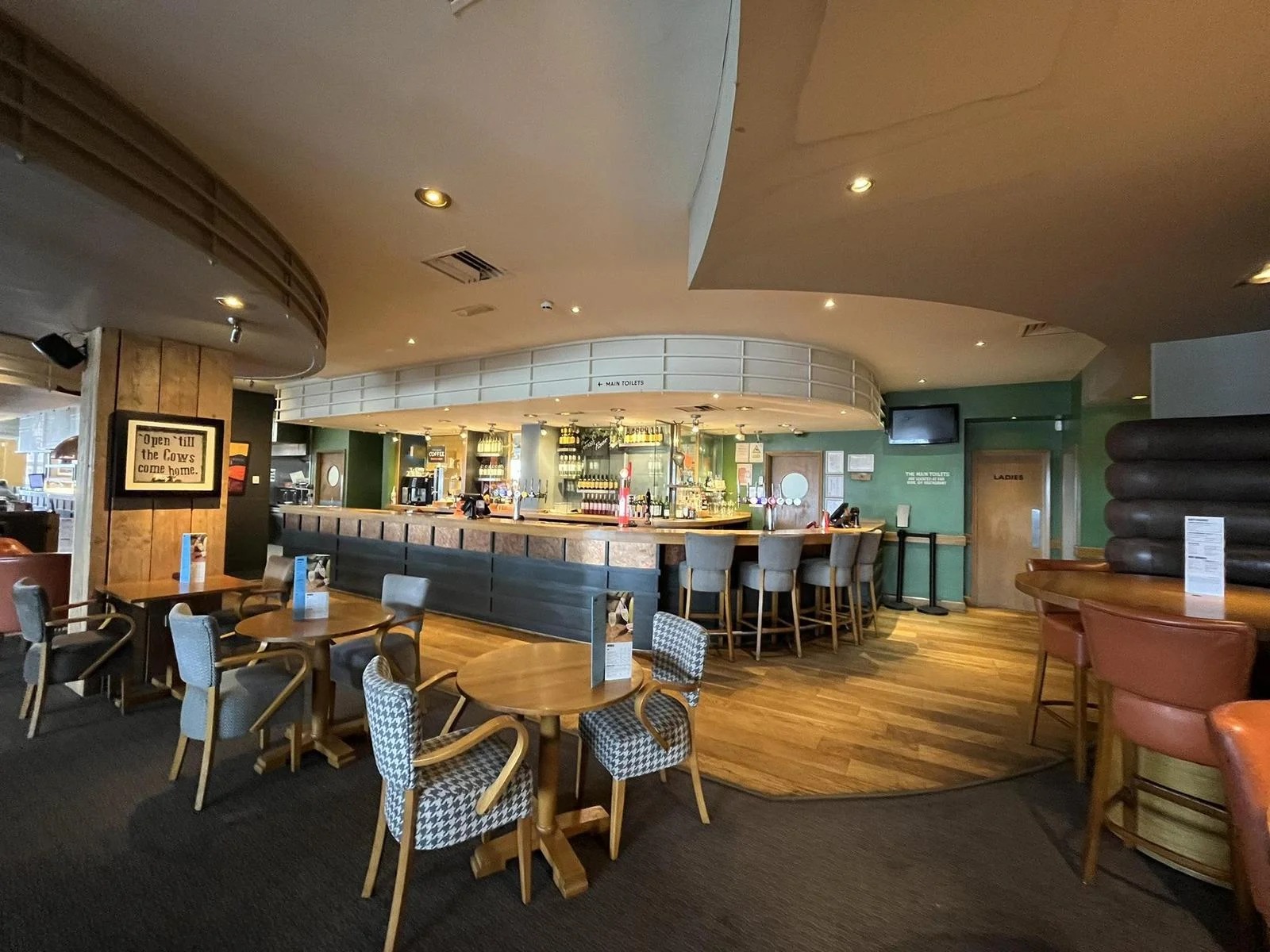 A long curved bar with stools, tables and chairs, and a framed sign reading Open till the cows come home.