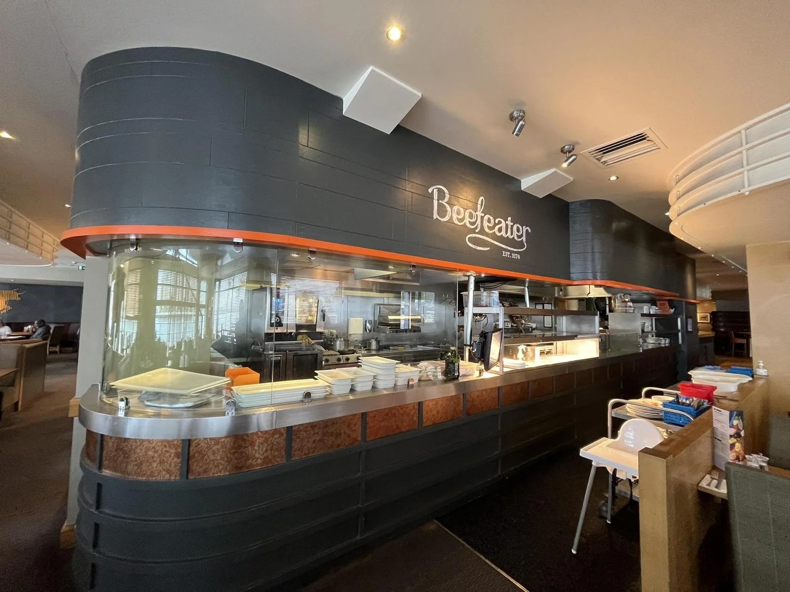 An open grill pass with a stainless steel service counter beneath a Beefeater sign reading est. 1974.