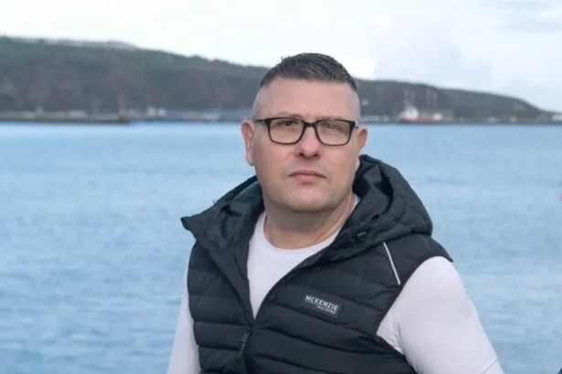 Brian Buckle, a man wearing glasses and a black gilet over a white top, standing in front of Fishguard harbour