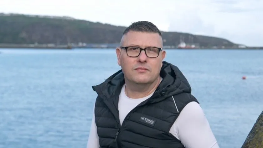 Brian Buckle, a man wearing glasses and a black gilet over a white top, standing in front of Fishguard harbour