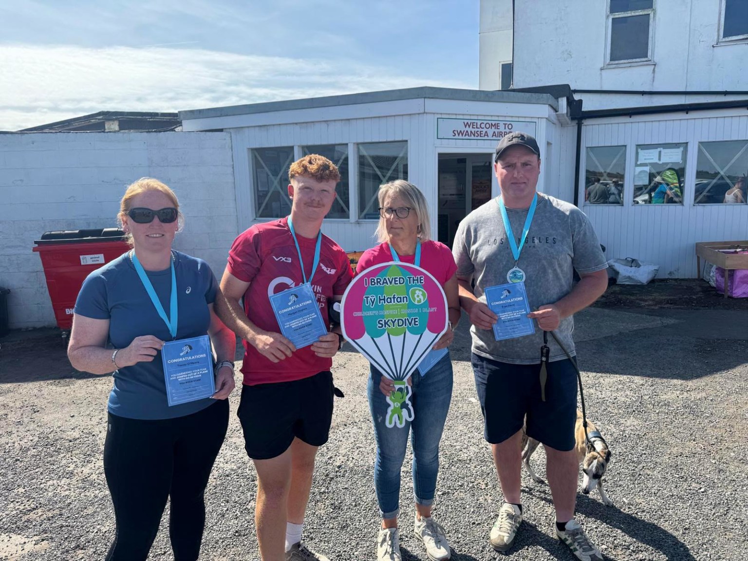 Four people holding skydive certificates outside the terminal at Swansea Airport, one holding a Tŷ Hafan parachute sign