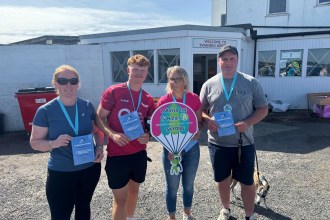 Four people holding skydive certificates outside the terminal at Swansea Airport, one holding a Tŷ Hafan parachute sign
