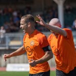 Pure Swansea captain Jordan Hughes celebrating a goal in an orange shirt, with team-mates joining him and a crowd behind
