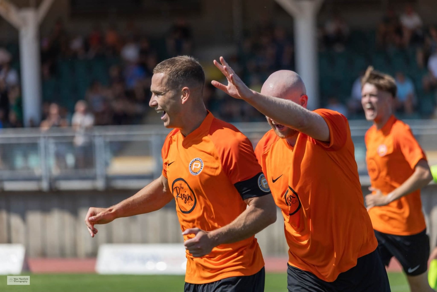 Pure Swansea captain Jordan Hughes celebrating a goal in an orange shirt, with team-mates joining him and a crowd behind