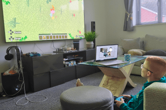 A boy sitting with a controller playing Minecraft on a large screen, with a laptop video call beside him