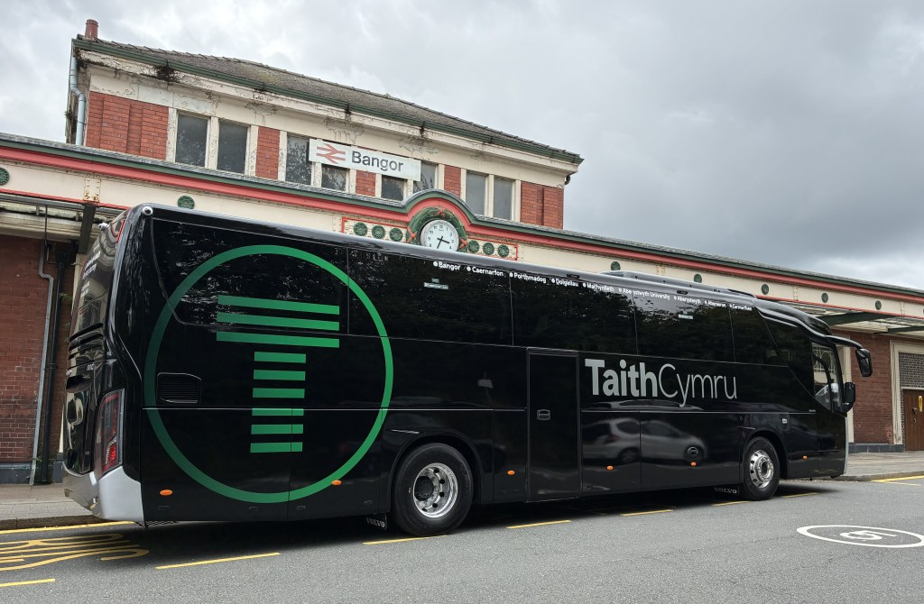 A black TaithCymru coach with a green logo parked outside Bangor railway station, its side listing the route's stops from Bangor to Carmarthen