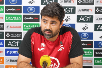 Swansea City head coach Vitor Matos speaking at a press conference in front of a sponsors' backdrop