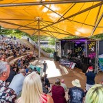 Audience on the stepped seating at Swansea amphitheatre beneath a yellow sail canopy, watching a band on the outdoor stage