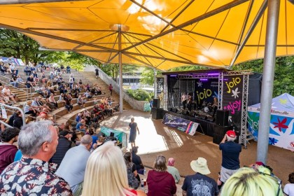 Audience on the stepped seating at Swansea amphitheatre beneath a yellow sail canopy, watching a band on the outdoor stage