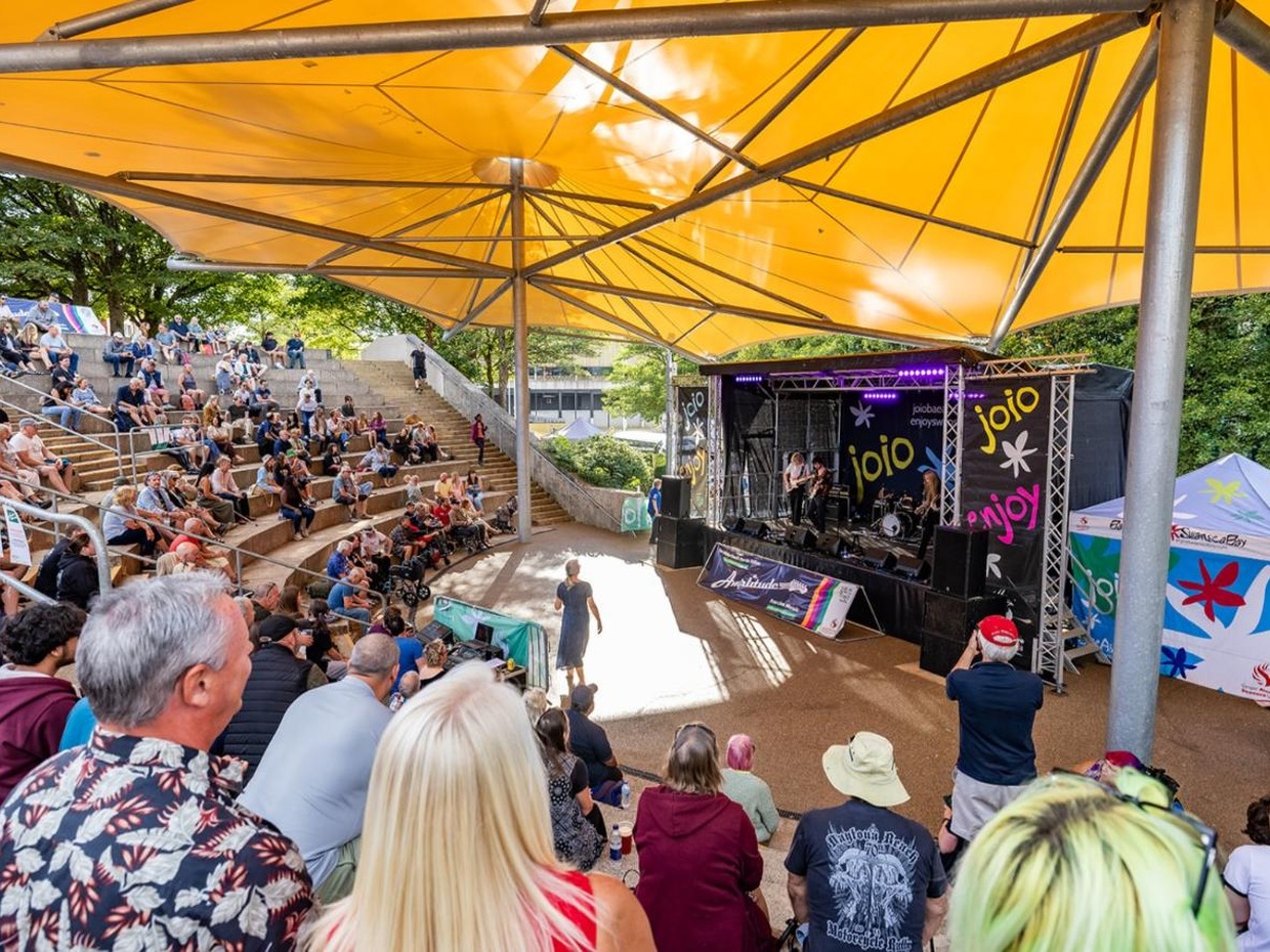 Audience on the stepped seating at Swansea amphitheatre beneath a yellow sail canopy, watching a band on the outdoor stage