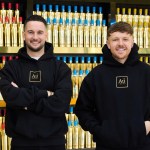 Two men in black Au Vodka hoodies standing in front of shelves stacked with gold vodka bottles