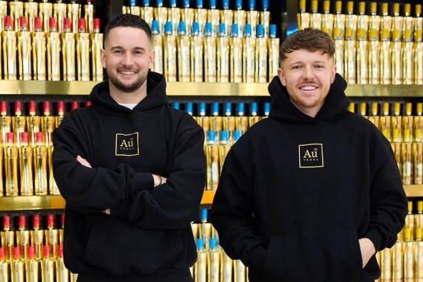 Two men in black Au Vodka hoodies standing in front of shelves stacked with gold vodka bottles