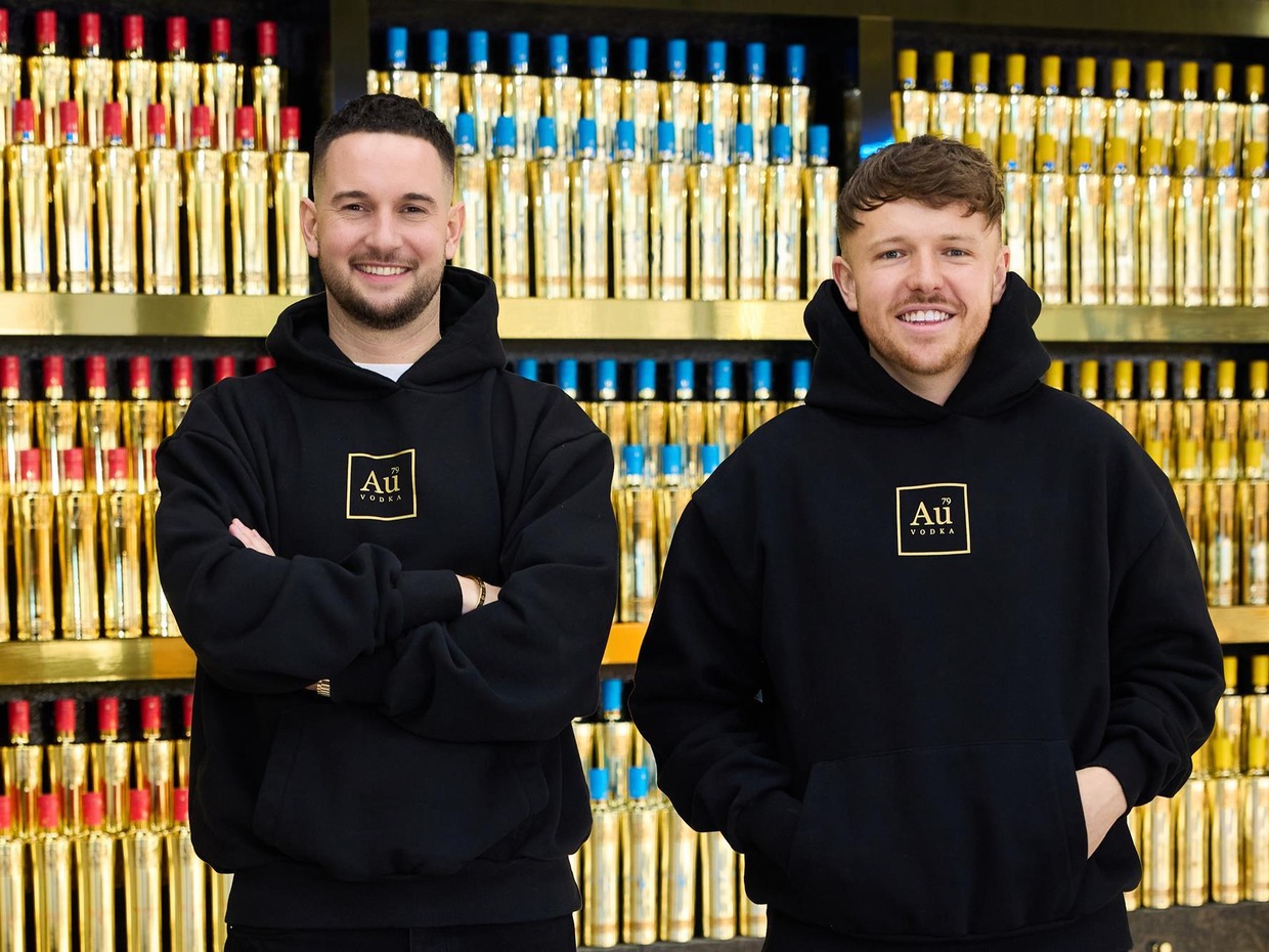 Two men in black Au Vodka hoodies standing in front of shelves stacked with gold vodka bottles