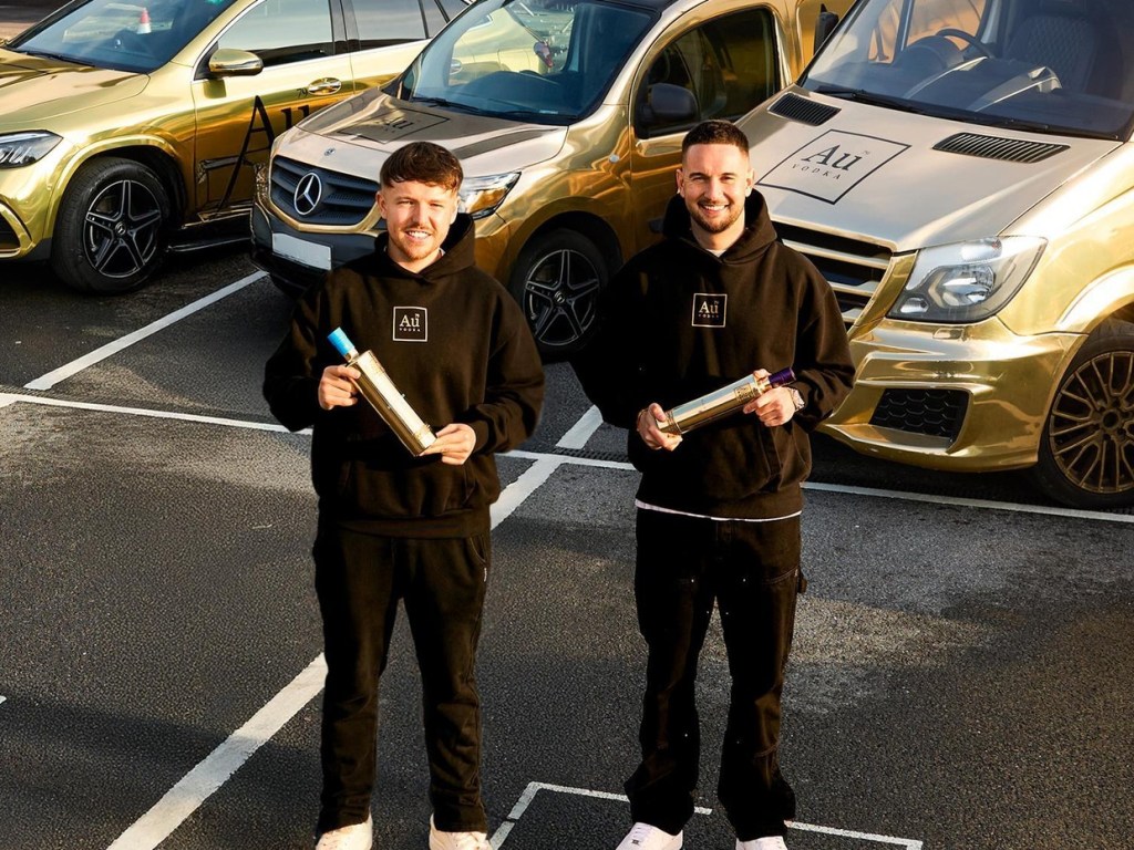 Two men in black Au Vodka hoodies holding gold bottles in front of three gold-wrapped Mercedes vehicles