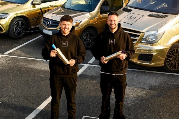 Two men in black Au Vodka hoodies holding gold bottles in front of three gold-wrapped Mercedes vehicles
