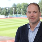 Alun Wyn Jones in a jacket and open-necked shirt, standing beside a running track and pitch