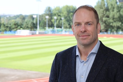 Alun Wyn Jones in a jacket and open-necked shirt, standing beside a running track and pitch