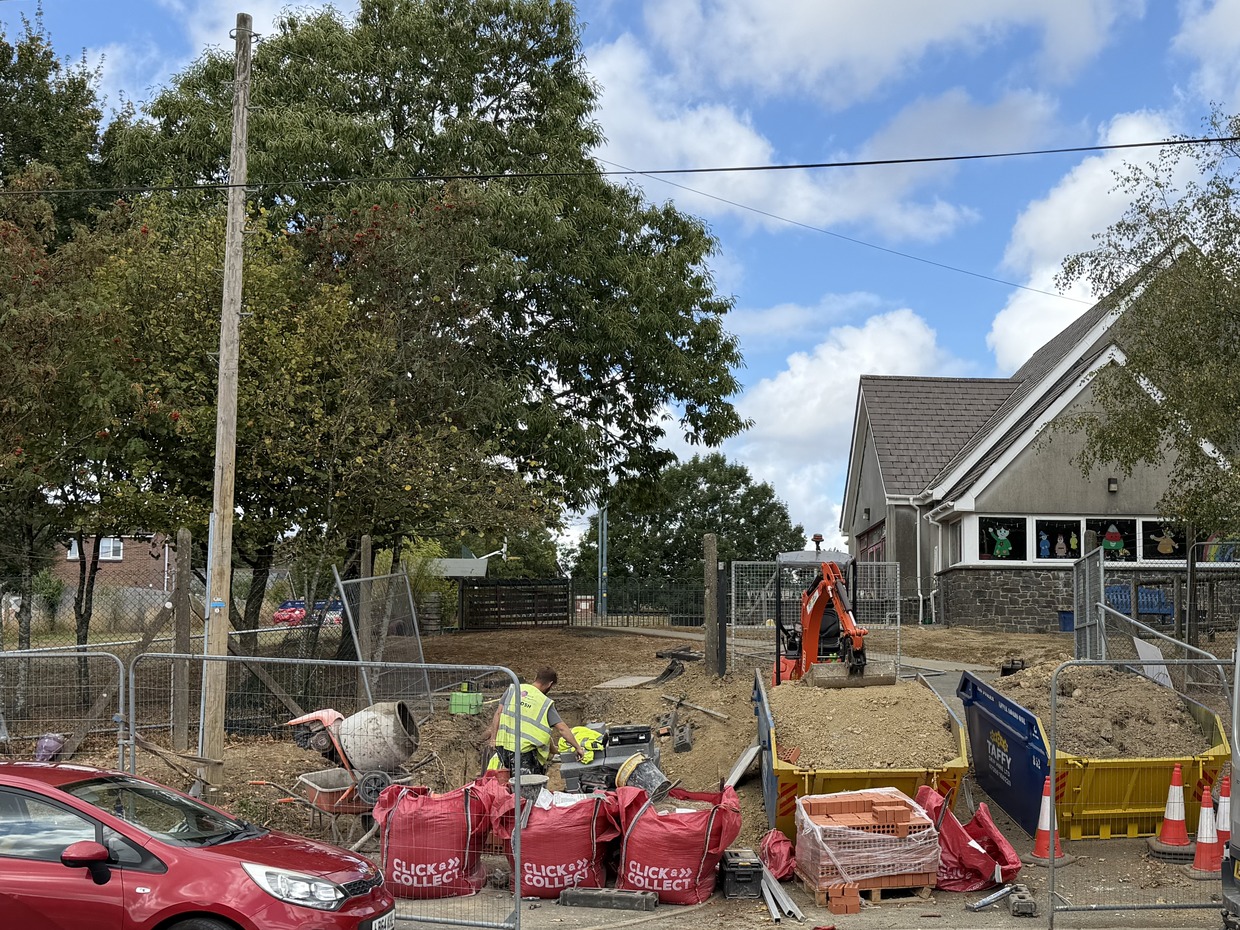 Metal fencing, a cement mixer, bulk bags and a mini digger on bare ground at the entrance to Ysgol Bancyfelin