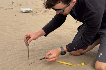 A man kneels on a beach holding a thermometer probe in the sand next to a disposable barbecue tray