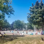 Families in and around the pool at Blackpill Lido on a sunny day, with splash fountains, swings and trees around the water