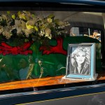 Close view through a hearse window showing a coffin draped in the Welsh flag, white lilies and roses laid on top, and a framed black and white portrait of Bonnie Tyler