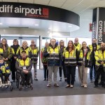 A group of Cardiff Airport assistance staff and service users, including wheelchair users, in the terminal beneath the Cardiff Airport sign