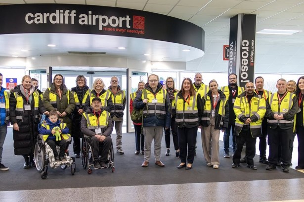 A group of Cardiff Airport assistance staff and service users, including wheelchair users, in the terminal beneath the Cardiff Airport sign