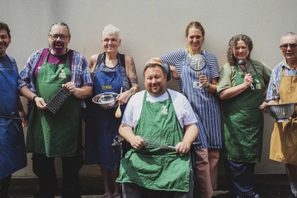 Seven Cegin Llanelli volunteer cooks in aprons holding kitchen utensils, lined up against a wall, one seated in a wheelchair holding a whisk