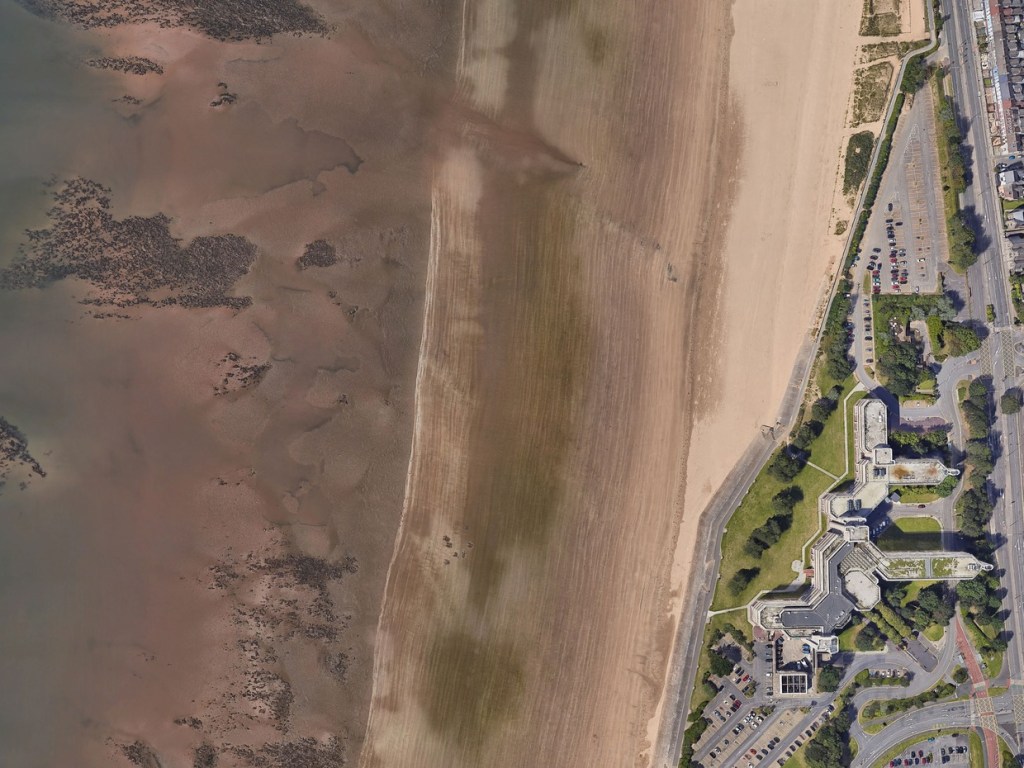 Overhead satellite view of Swansea beach at low tide, with the Civic Centre and its car parks along the top of the sand