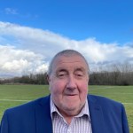 Cllr Peter Cooper, wearing a navy jacket and checked shirt, standing in front of playing fields under a blue sky