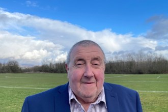 Cllr Peter Cooper, wearing a navy jacket and checked shirt, standing in front of playing fields under a blue sky