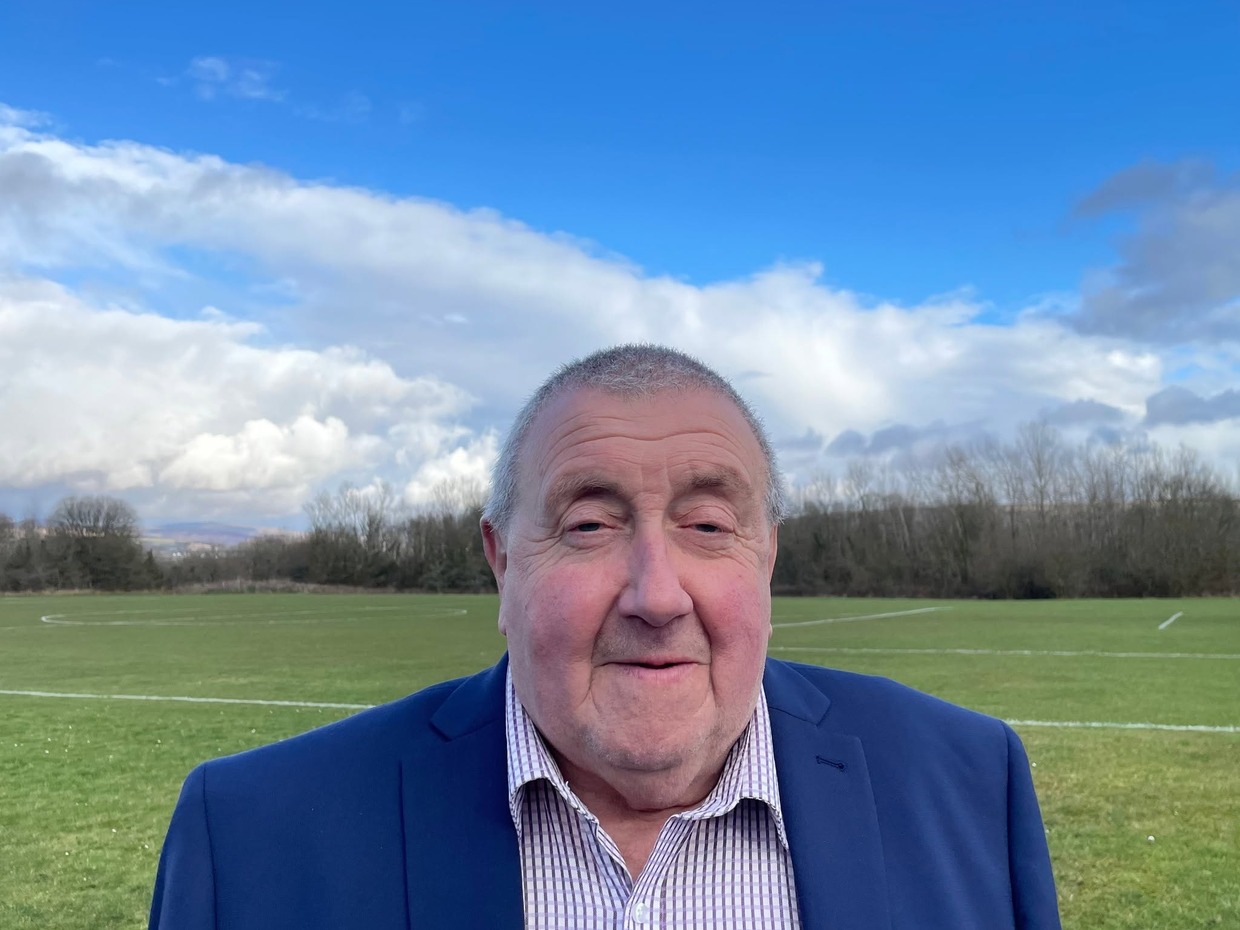 Cllr Peter Cooper, wearing a navy jacket and checked shirt, standing in front of playing fields under a blue sky