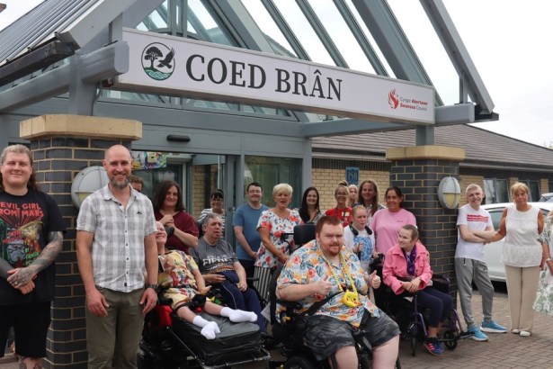 A group of Coed Brân service users and staff, including wheelchair users, beneath the Coed Brân sign at the Llansamlet centre