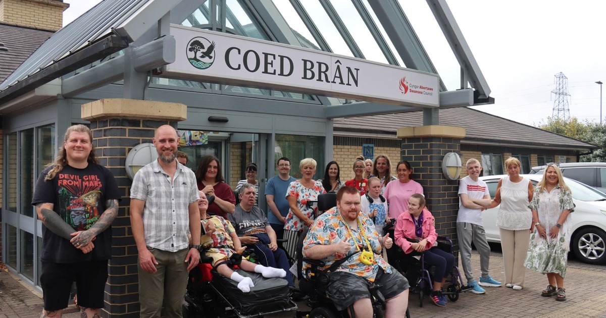A group of Coed Brân service users and staff, including wheelchair users, beneath the Coed Brân sign at the Llansamlet centre