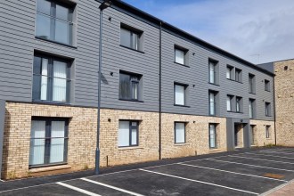 A completed five-storey block at the County Flats in Aberavon, with pale brick, grey cladding and a corner tower, under a blue sky