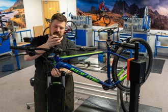 Chris Dower working on a blue mountain bike held in a repair stand in the Cycle CPD training workshop, with benches and bikes behind him