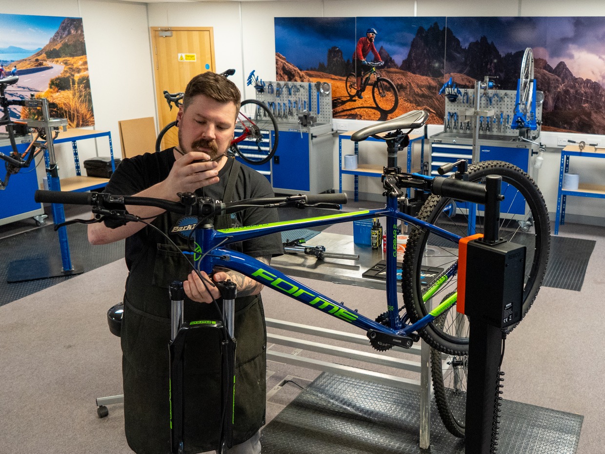 Chris Dower working on a blue mountain bike held in a repair stand in the Cycle CPD training workshop, with benches and bikes behind him