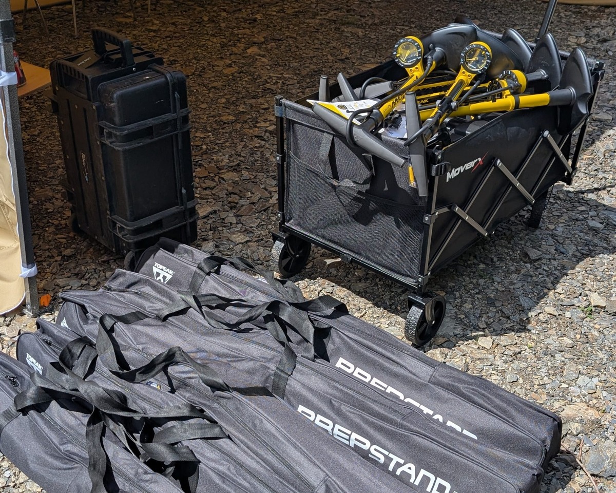 Folded portable bike work stands in carry bags, a tool case and a trolley loaded with yellow track pumps on a gravel yard