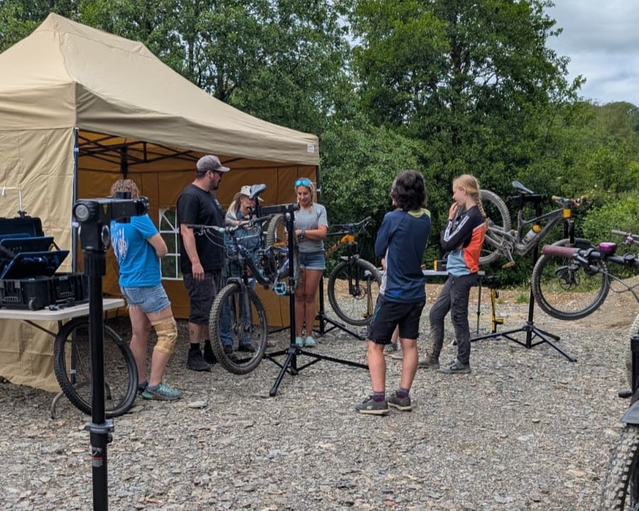 Riders gathered around portable bike work stands beside a gazebo at a mountain bike centre, with mountain bikes lined up behind