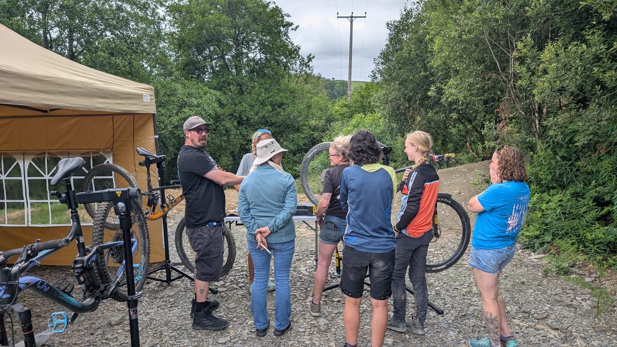 A group of riders standing around bikes held in portable work stands on a gravel yard beside a gazebo, listening to a trainer