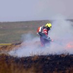 Two firefighters in red wildfire kit working in smoke on burning moorland, one with a shovel and one using a leaf blower against flames