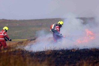 Two firefighters in red wildfire kit working in smoke on burning moorland, one with a shovel and one using a leaf blower against flames