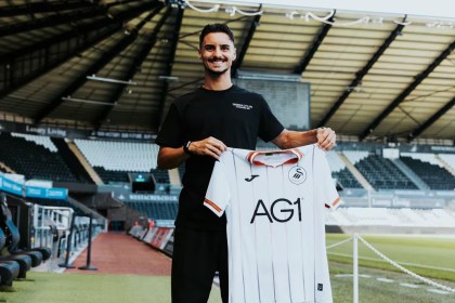 Stephen Eustáquio smiling and holding up a white Swansea City home shirt pitchside at the Swansea.com Stadium