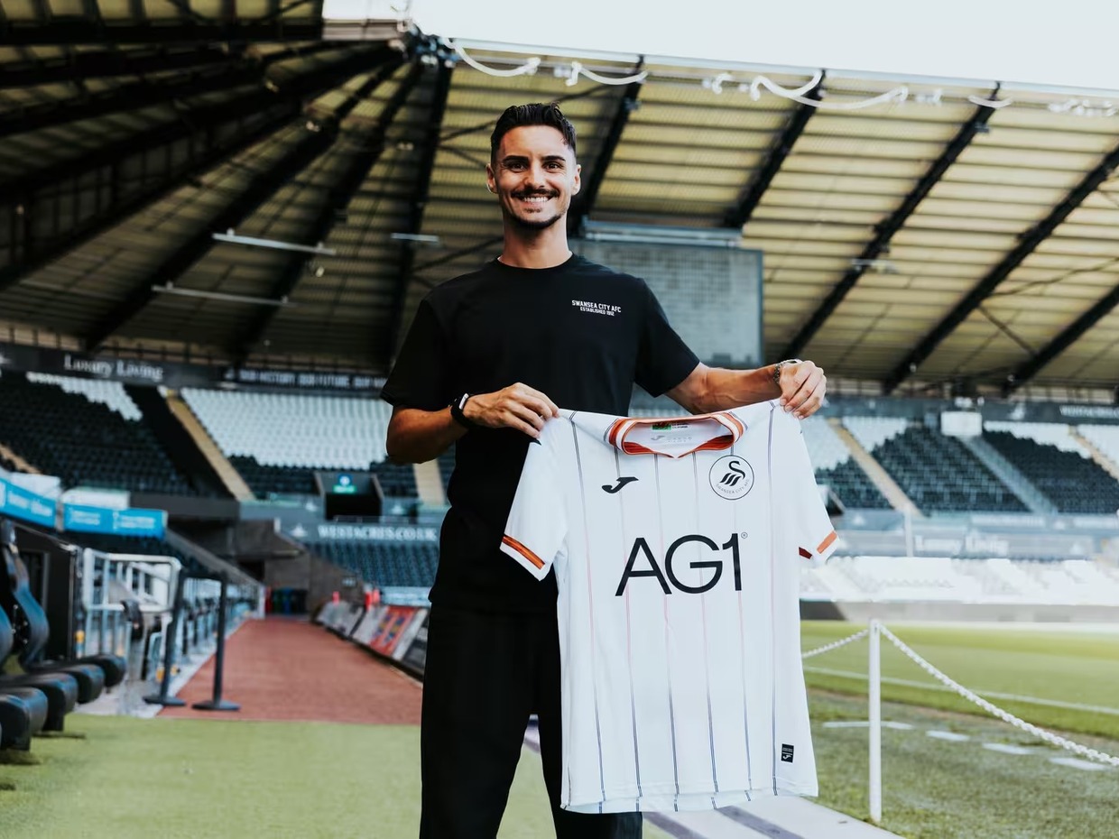 Stephen Eustáquio smiling and holding up a white Swansea City home shirt pitchside at the Swansea.com Stadium