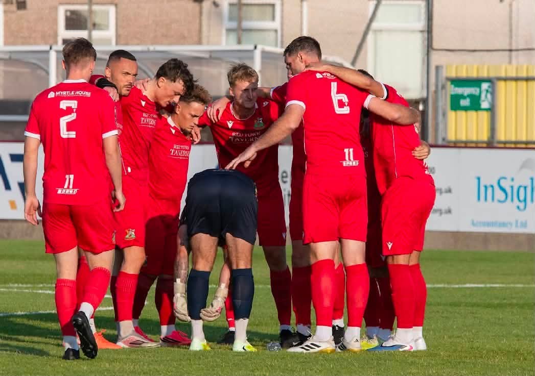 Llanelli Town players in red shirts gather in a huddle on the pitch at Stebonheath before kick-off